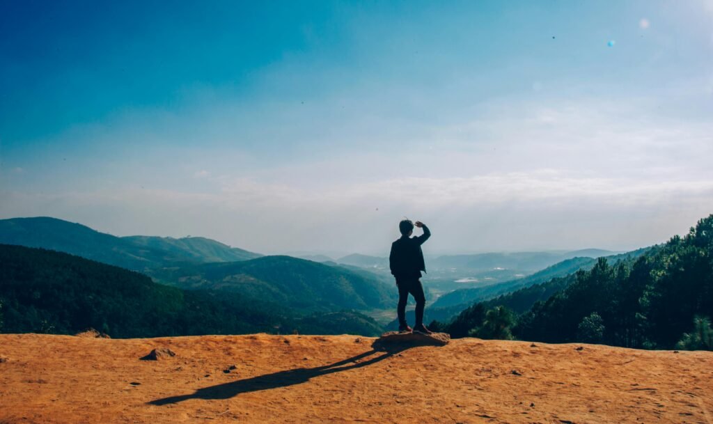 A lone traveler stands on a hilltop, gazing over a vast mountain landscape under a clear blue sky. motivated and happy